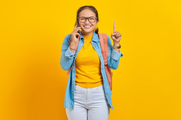 Portrait of cheerful young Asian woman student in casual clothes with backpack talking on mobile phone and pointing finger up, creating genius solutions isolated on yellow background