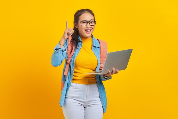 Портрет веселой молодой азиатской студентки в повседневной одежде с рюкзаком, использующей ноутбук и имеющей творческую идею, изолированную на желтом фоне. Образование в концепции университетского колледжа