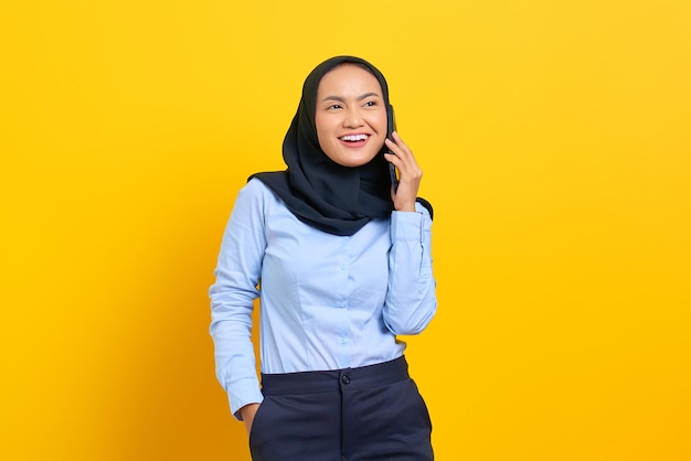 黄色の背景で隔離の携帯電話で話している陽気な若いアジアの女性の肖像画