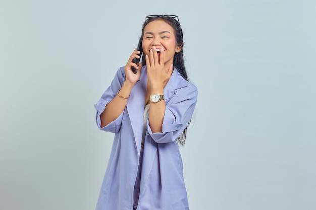 Portrait of cheerful young Asian woman talking on mobile phone with hands covering mouth isolated on white background