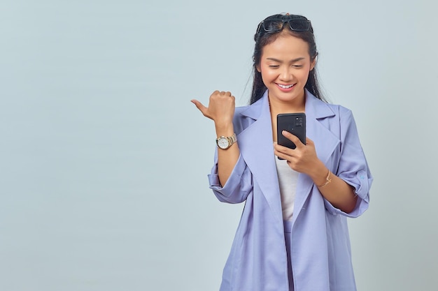 Portrait of cheerful young Asian woman using mobile phone and pointing to copy space isolated on white background