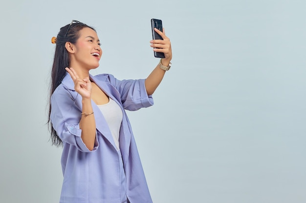 Портрет веселой молодой азиатки, использующей мобильный телефон, делает селфи, делает знак мира на белом фоне