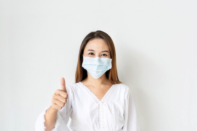 Portrait of cheerful young Asian woman with protective face mask thumb up posing and smiling at camera