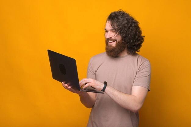 Portrait of cheerful young bearded man with long curly hair using laptop over yellow