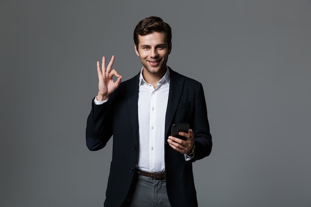 Portrait of a cheerful young businessman dressed in suit isolated over gray wall, holding mobile phone, showing ok gesture