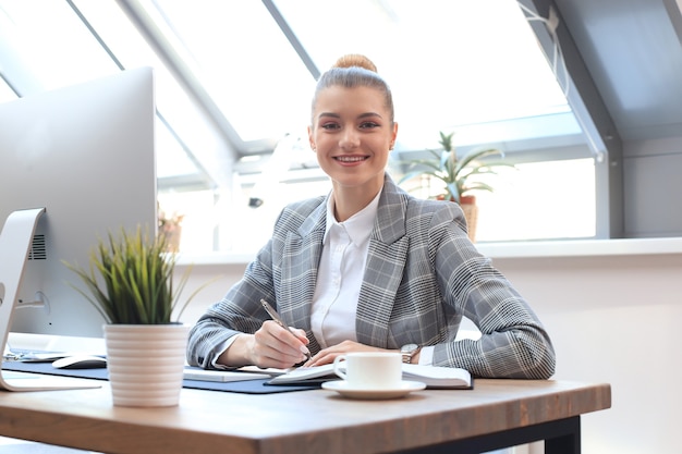 portrait of a cheerful young businesswoman sitting at the table in office and looking at camera.