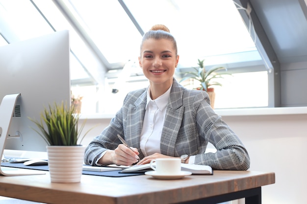 Portrait of a cheerful young businesswoman sitting at the table in office and looking at camera.