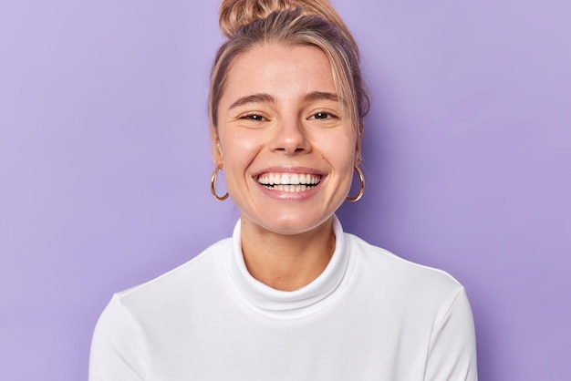 Portrait of cheerful young female model smiles broadly shows perfect teeth wears golden earrings and casual white jumper looks gladfully poses against purple background Happy emoions concept