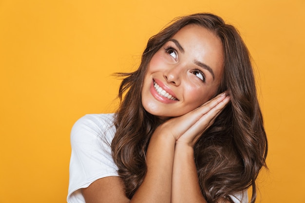 Portrait of a cheerful young girl with long brunette hair standing over yellow wall, holding folded arms at her cheek