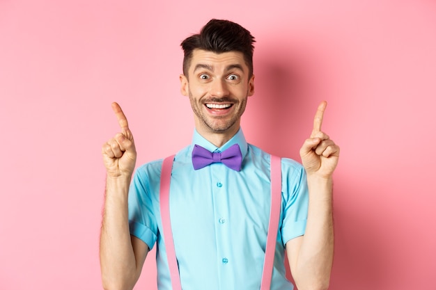 쾌활 한 젊은 남자 콧수염과 손가락을 가리키는 강모, 최고 로고를 보여주는 분홍색 배경에 서 카메라에 행복 미소의 초상화.