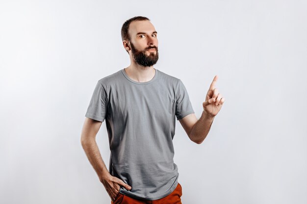 Portrait of cheerful young handsome man smiling looking at camera pointing finger upwards on white background with space for advertising mock up