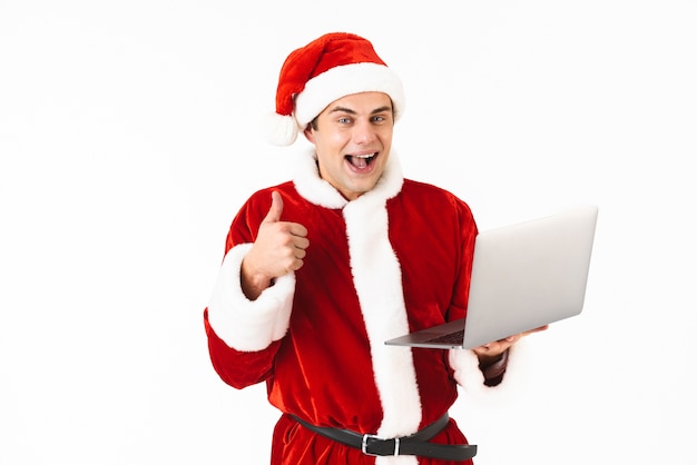 Portrait of a cheerful young man dressed in santa claus costume standing isolated over white space, holding laptop computer