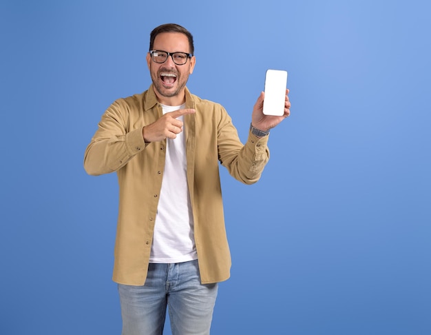 Foto ritratto di un giovane allegro che indica uno smartphone e un'app di appuntamenti pubblicitaria su sfondo blu