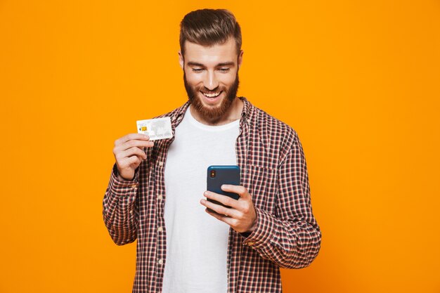 Portrait of a cheerful young man wearing casual clothes holding mobile phone showing plastic credit card