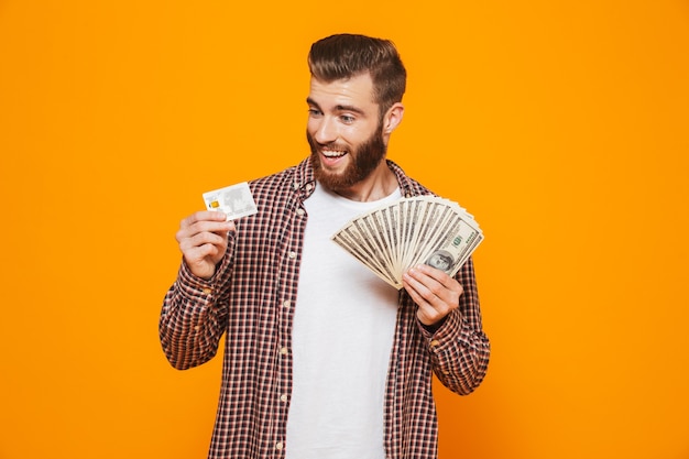 Portrait of a cheerful young man wearing casual clothes holding money banknotes showing plastic credit card