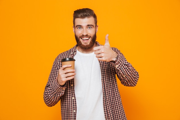 Portrait of a cheerful young man wearing casual clothes holding takeaway coffee giving thumbs up
