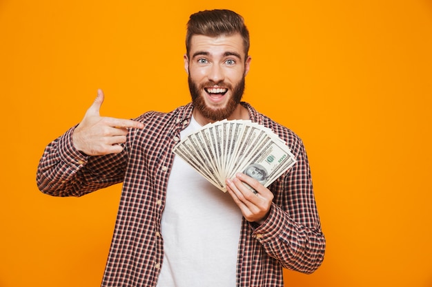 Portrait of a cheerful young man wearing casual clothes showing money banknotes pointing finger