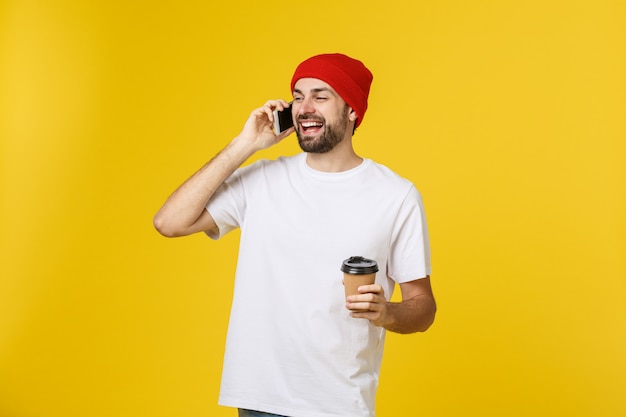 Portrait of a cheerful young man wearing casual clothes standing isolated over yellow, holding mobile phone, drinking takeaway coffee