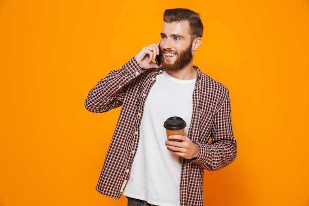 Portrait of a cheerful young man wearing casual clothes talking on mobile phone drinking takeaway coffee