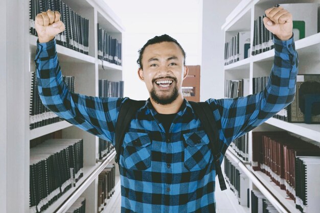 Portrait of cheerful young man with arms raised standing in library