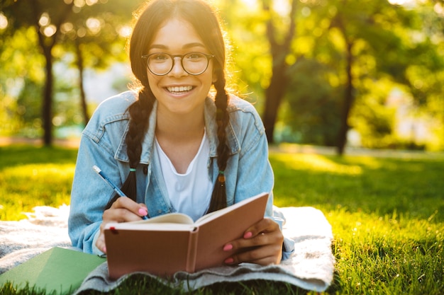 Portrait of a cheerful young student girl wearing eyeglasses sitting outdoors in nature park writing notes reading book. Looking camera.