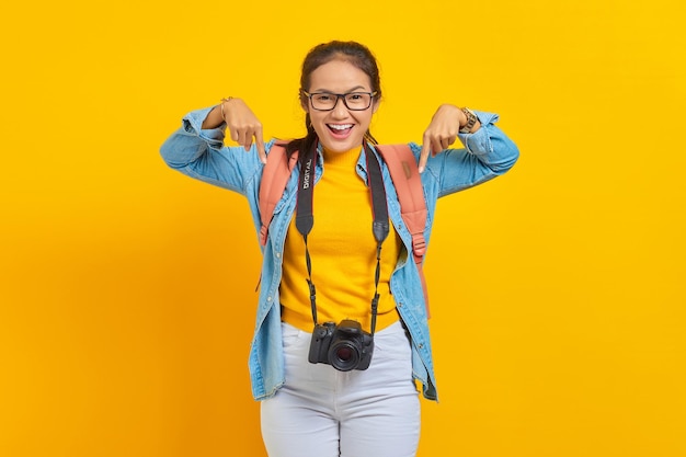 Portrait of cheerful young traveler Asian woman with backpack and camera in denim clothes while pointing at copy space with fingers isolated on yellow background. Air flight journey concept