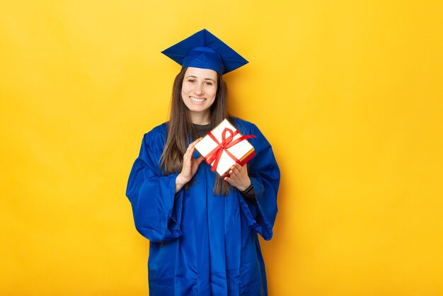 Portrait of cheerful young woman in blue robe graduating and holding gift box over yellow background