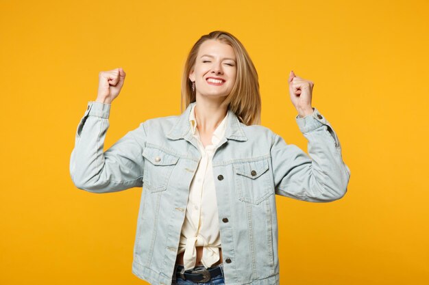 Portrait of cheerful young woman in denim casual clothes keeping eyes closed, doing winner gesture isolated on bright yellow orange background in studio. People lifestyle concept. Mock up copy space.