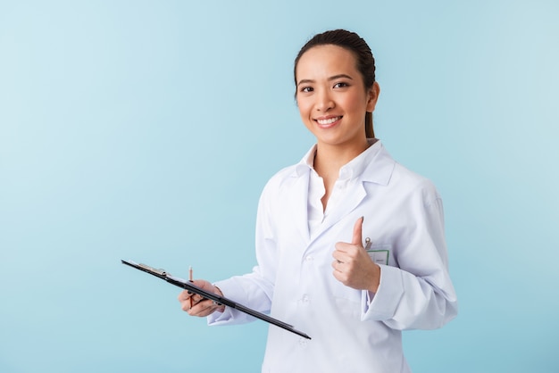 portrait of a cheerful young woman doctor posing isolated over blue wall holding clipboard showing thumbs up.