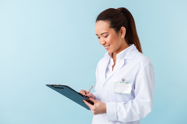 portrait of a cheerful young woman doctor posing isolated over blue wall holding clipboard.