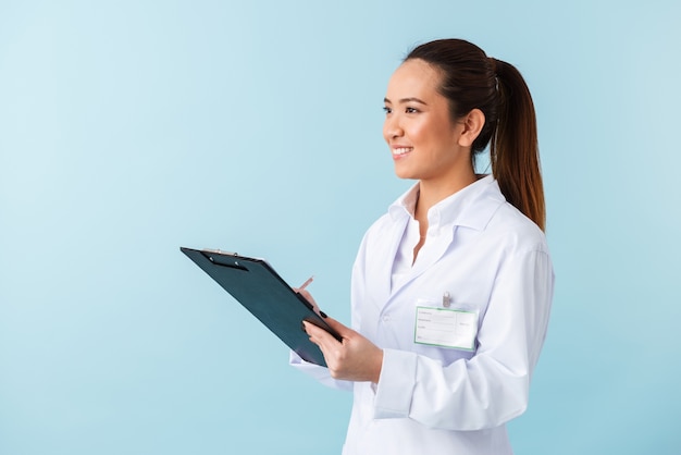 portrait of a cheerful young woman doctor posing isolated over blue wall holding clipboard.