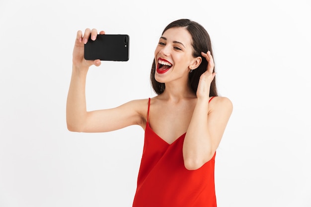 Photo portrait of a cheerful young woman in dress isolated, taking selfie with mobile phone