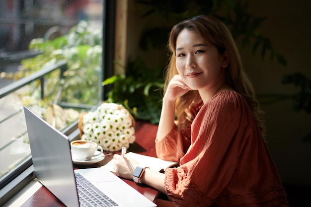 Portrait of cheerful young woman drinking coffee and studying in cafe