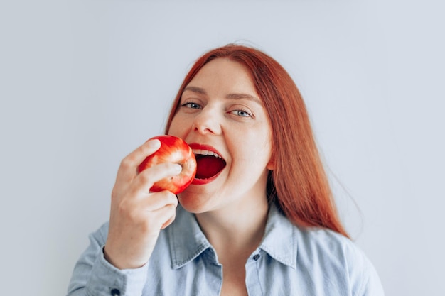 灰色の壁の背景に赤いリンゴを食べる陽気な若い女性の肖像画