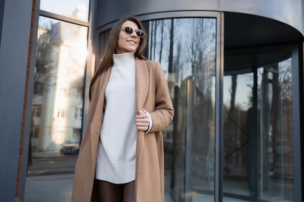 Portrait of cheerful young woman in glasses and brown coat outdoors