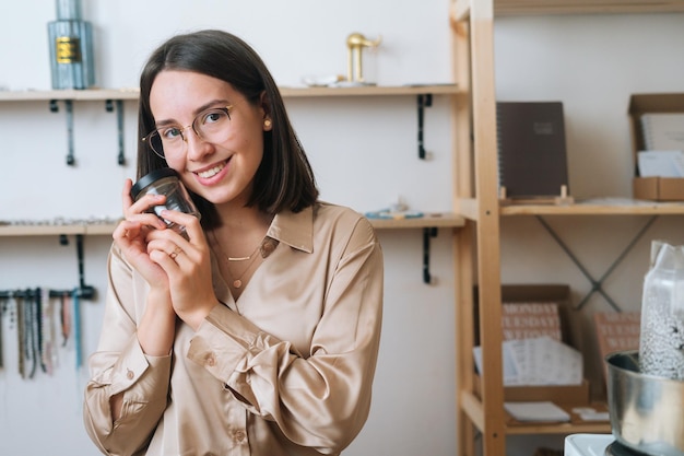 Portrait of cheerful young woman in glasses holding little empty transparent glass jar in hands sitting at table, looking at camera. Process of making handmade natural candle at workshop.