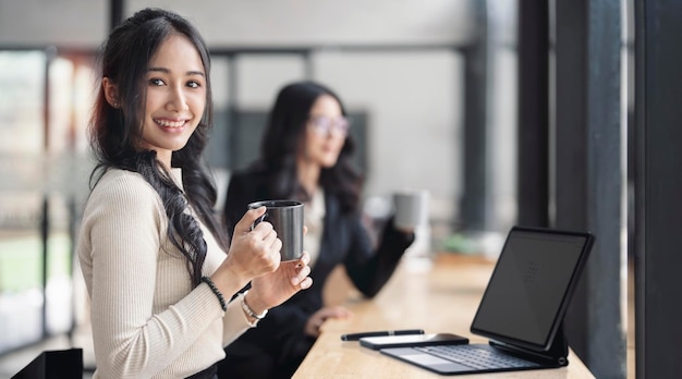 Portrait of cheerful young woman and her friend or colleague relaxing with coffee cup during business meeting at cafe smiling and looking at camera