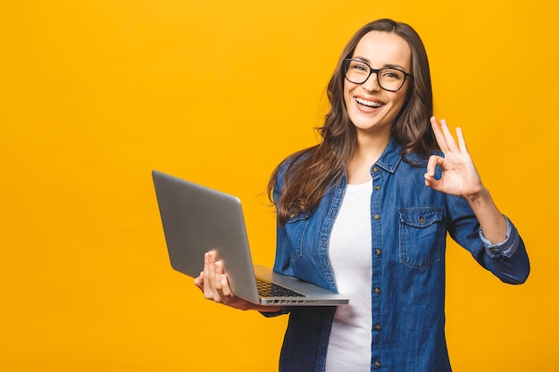 Portrait of a cheerful young woman holding laptop computer and showing ok