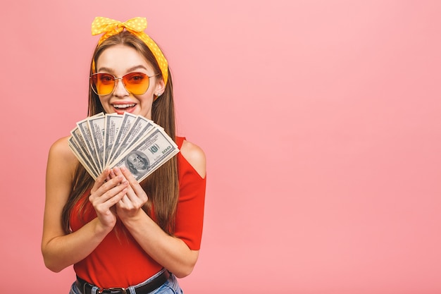 Photo portrait of a cheerful young woman holding money banknotes and celebrating isolated over pink background