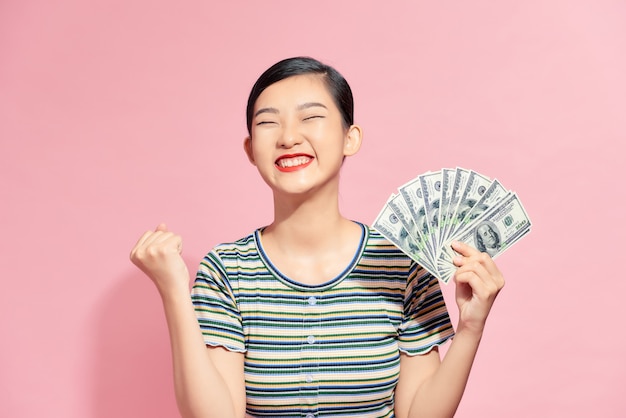 Portrait of a cheerful young woman holding money banknotes and celebrating isolated over pink background