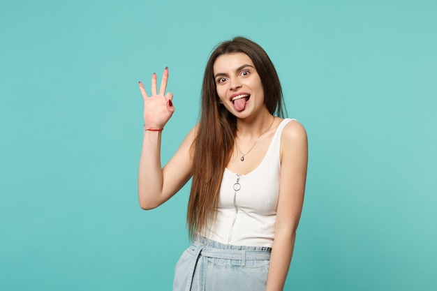 Portrait of cheerful young woman in light casual clothes showing tongue, OK gesture isolated on blue turquoise wall background in studio. People sincere emotions lifestyle concept. Mock up copy space.