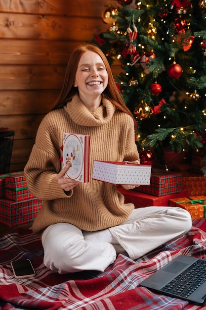 Portrait of cheerful young woman opening gift box with christmas present on background of xmas tree