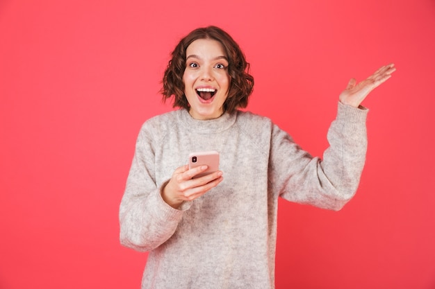 Portrait of a cheerful young woman standing isolated over pink, holding mobile phone