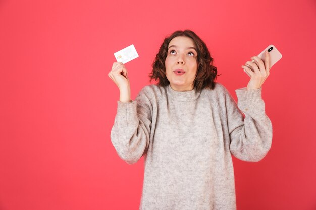 ピンクの上に孤立して立っている陽気な若い女性の肖像画、携帯電話を使用して、プラスチックのクレジットカードを示しています