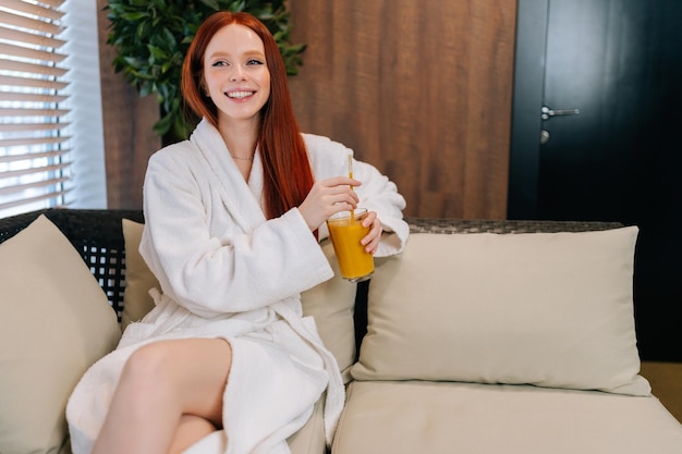 Portrait of cheerful young woman wearing white bathrobe enjoying drinking cocktail sitting on sofa by window at spa resort Charming redhead female relaxing
