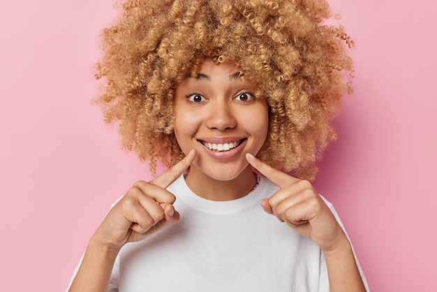 Ritratto di giovane donna allegra con i capelli ricci punta il dito indice al sorriso a trentadue denti essendo di buon umore vestito casualmente isolato su sfondo rosa concetto di persone ed emozioni positive