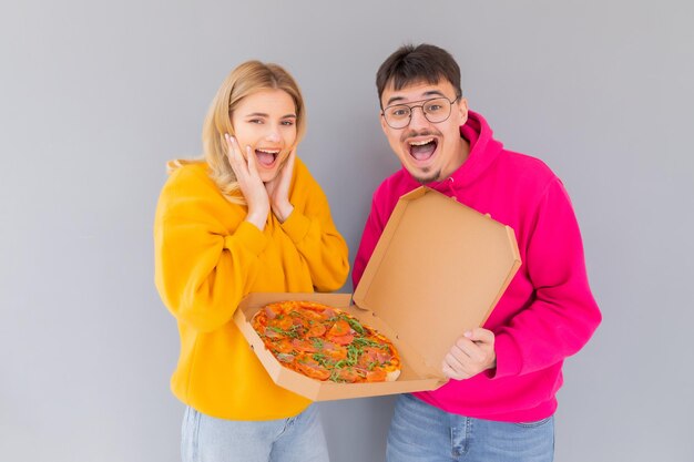 Portrait of cheery couple man and woman in colored sweaters smiling while eating pizza