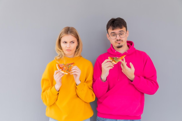 Portrait of cheery couple man and woman in colored sweaters smiling while eating pizza