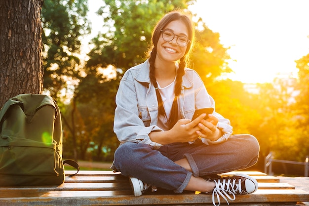 Portrait of a cheery cute young student girl wearing eyeglasses sitting on bench outdoors in nature park with beautiful sunlight using mobile phone