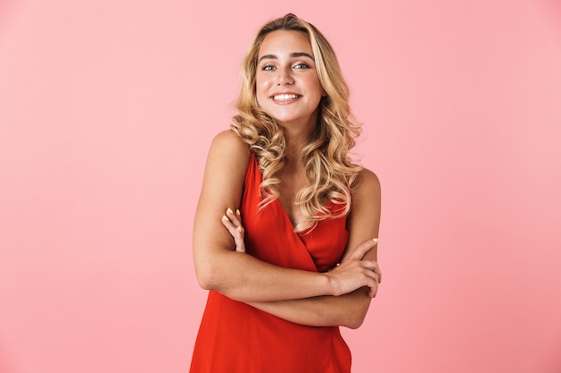 Portrait of a cheery smiling young pretty blonde cute woman in dress posing isolated over pink wall with arms crossed